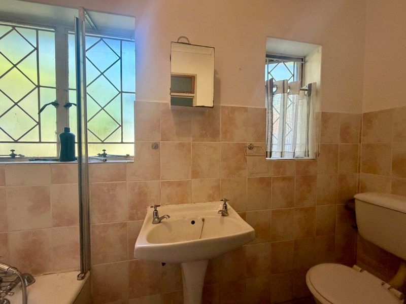 A dated bathroom featuring a pedestal basin, toilet, and shower area with tiled walls and natural light from two windows.