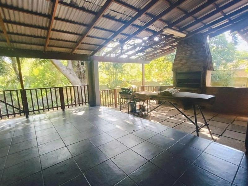 Covered outdoor patio featuring a built-in braai, tiled flooring, and timber roof structure, overlooking the garden area.