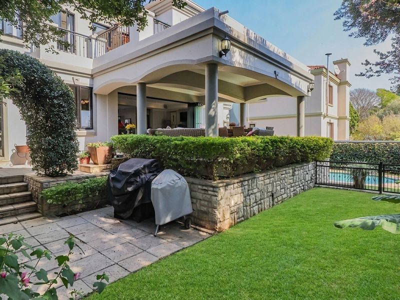 A well-maintained rear exterior featuring a covered patio, stone retaining wall, paved terrace, and a landscaped garden with a swimming pool.