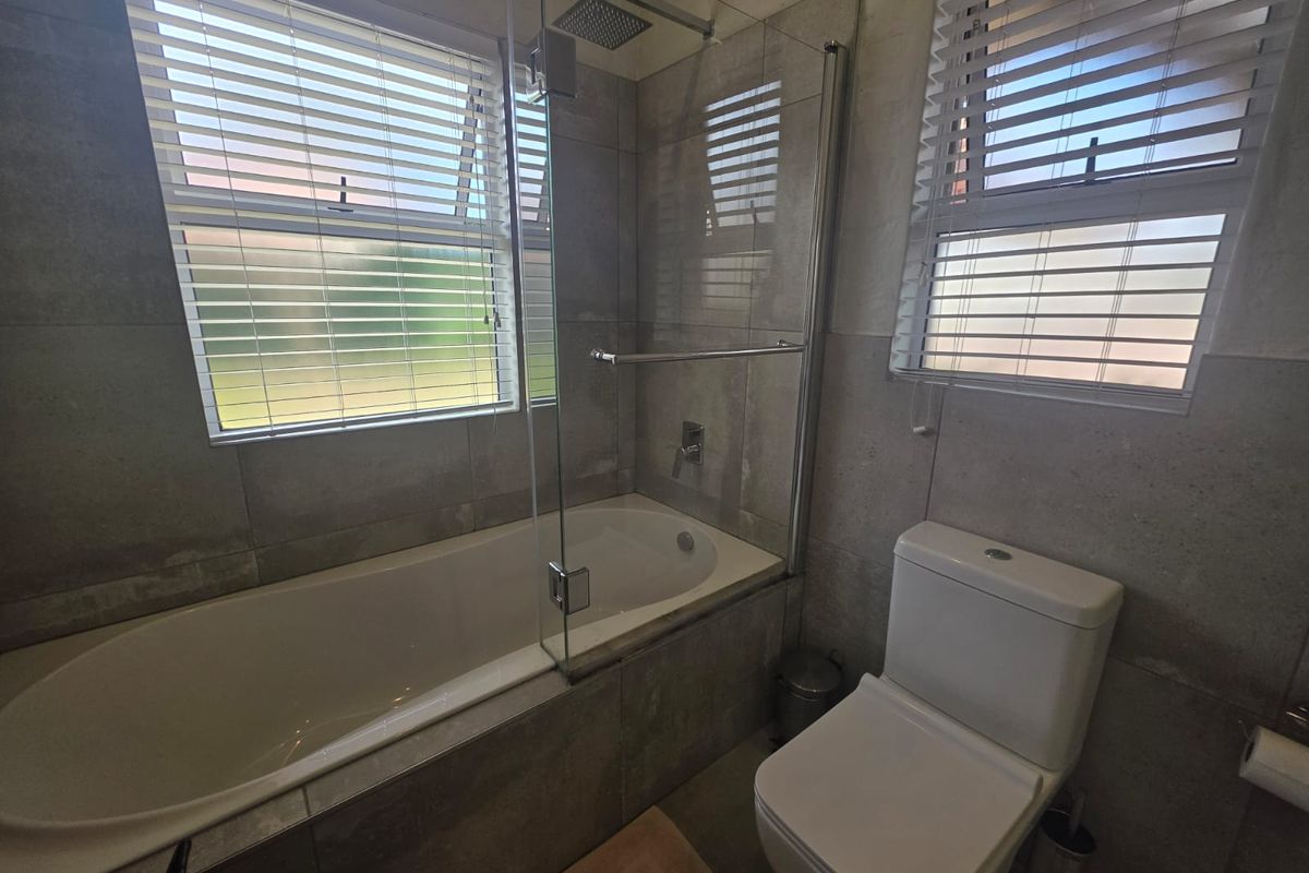 Modern bathroom featuring a built-in bathtub with an overhead shower, glass screen, toilet, and neutral grey wall and floor tiling.