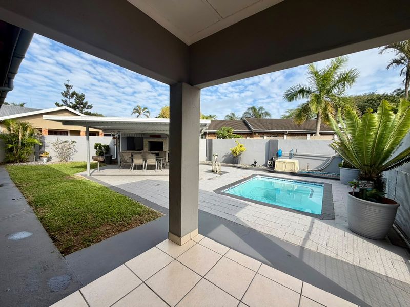 A spacious covered patio overlooking a paved courtyard with a swimming pool, built-in braai area, and well-maintained garden space.