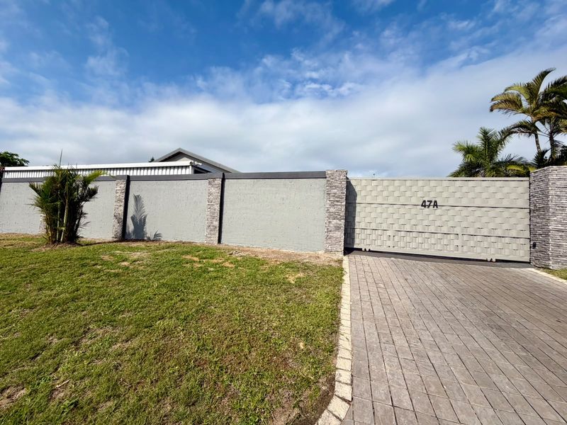 Front view of the property featuring a paved driveway, secure sliding gate, and a well-maintained boundary wall with stone-clad pillars.