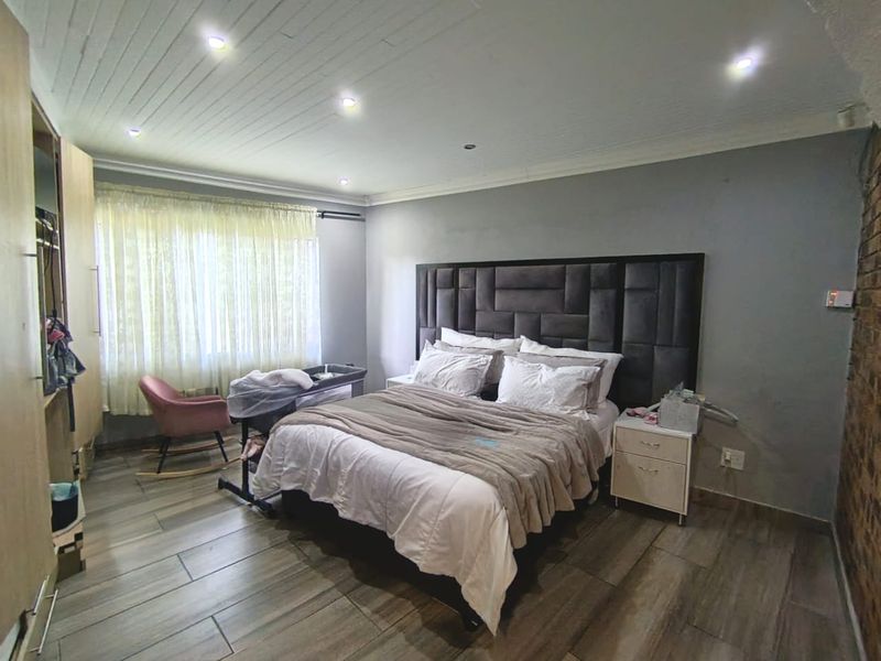 A spacious bedroom featuring timber-look floor tiles, recessed downlighting, a feature exposed brick wall, and built-in wardrobe storage.