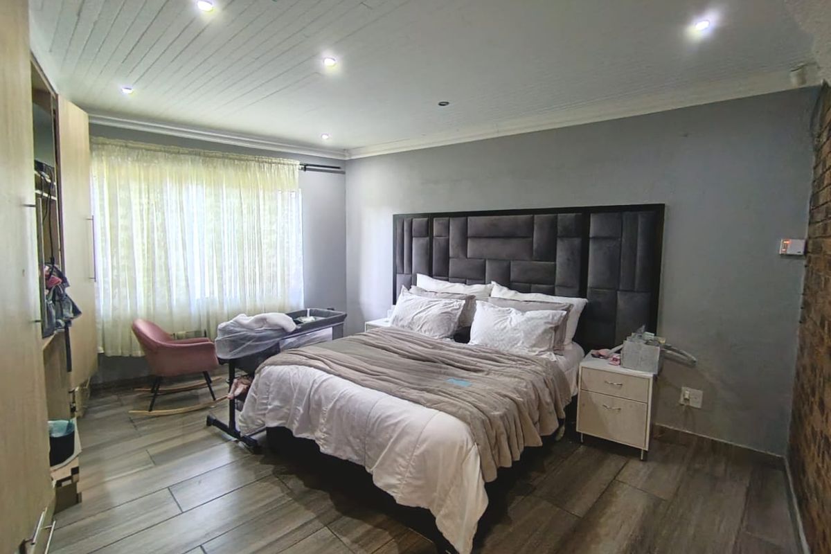 A spacious bedroom featuring timber-look floor tiles, recessed downlighting, a feature exposed brick wall, and built-in wardrobe storage.