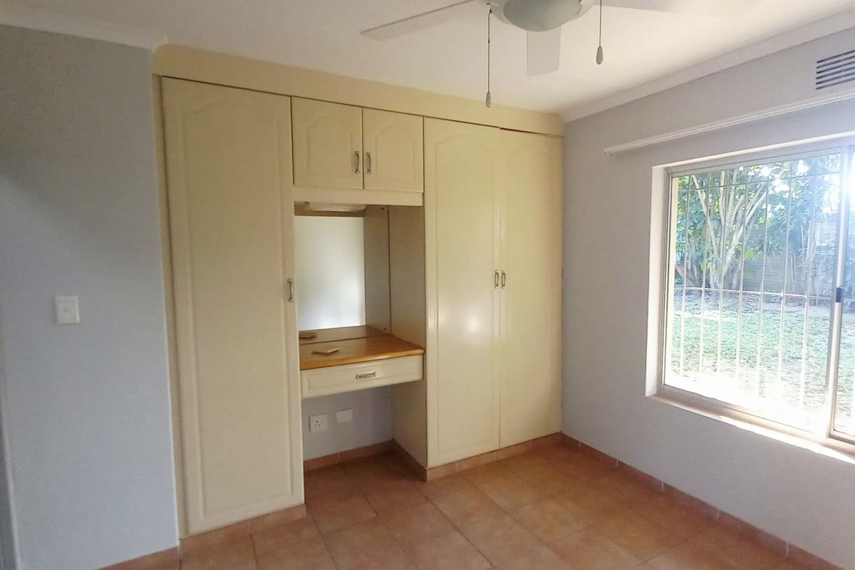 A bright bedroom featuring built-in cupboards with a central vanity nook, tiled flooring, and a ceiling fan, overlooking the garden through security-barred windows.