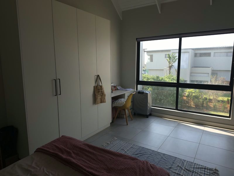 A bright bedroom featuring built-in cupboards, tiled flooring, and a large window with natural light, set under a vaulted ceiling.