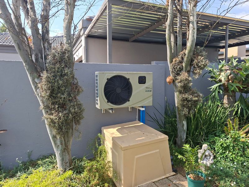 A rear exterior view showing a wall-mounted air conditioning condenser unit, a metal pergola structure, and a boundary wall in a landscaped garden setting.