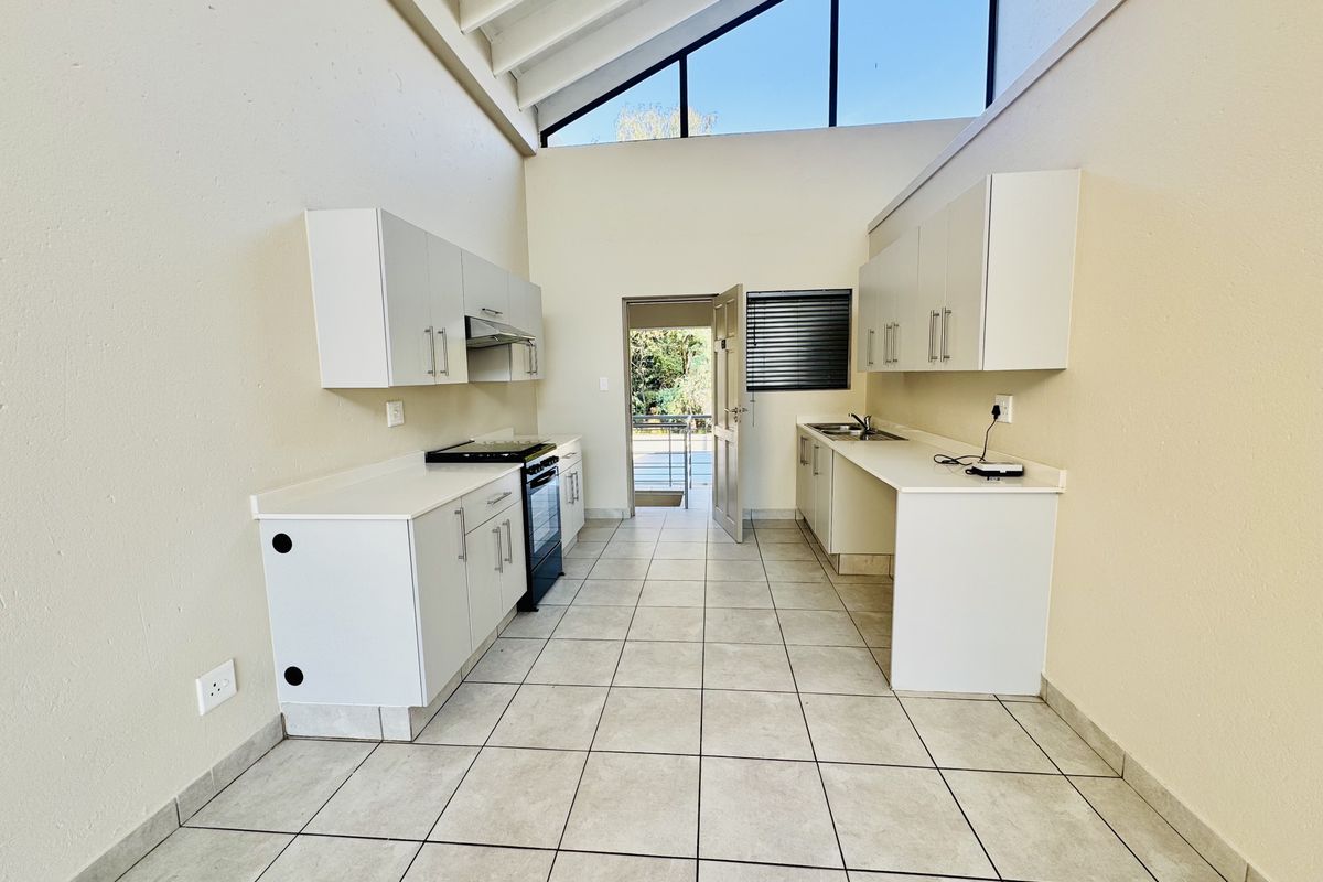 A bright, open-plan kitchen featuring white cabinetry, tiled flooring, and a high vaulted ceiling with clerestory windows, leading to an outdoor area.