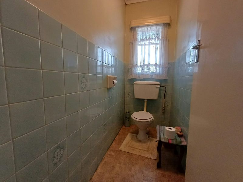 A compact bathroom featuring dated light blue ceramic wall tiles, a standard toilet, and a window with a lace curtain.