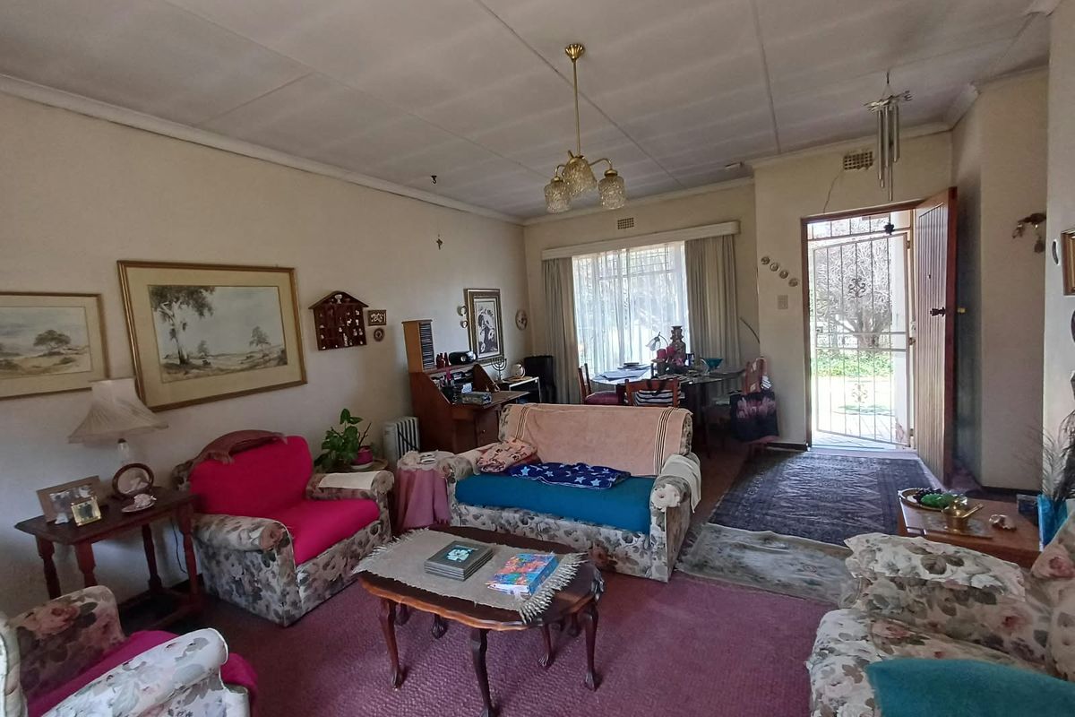 A spacious living room featuring carpeted flooring, a central pendant light, and direct access to the exterior through a security gate and timber door.