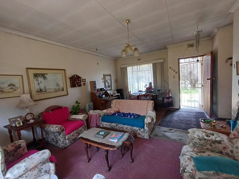 A spacious living room featuring carpeted flooring, a central pendant light, and direct access to the exterior through a security gate and timber door.