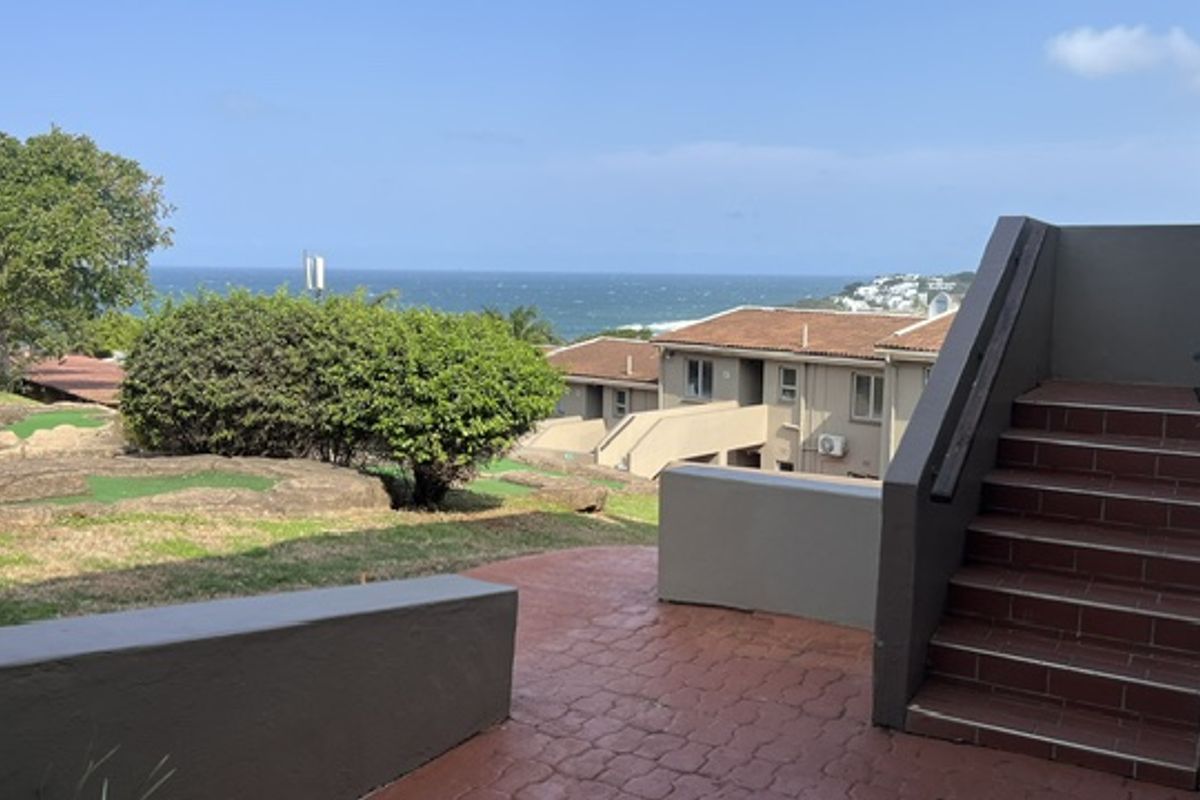 An outdoor rear patio area featuring a paved terrace, staircase, and retaining walls, offering scenic views of the surrounding complex and ocean.