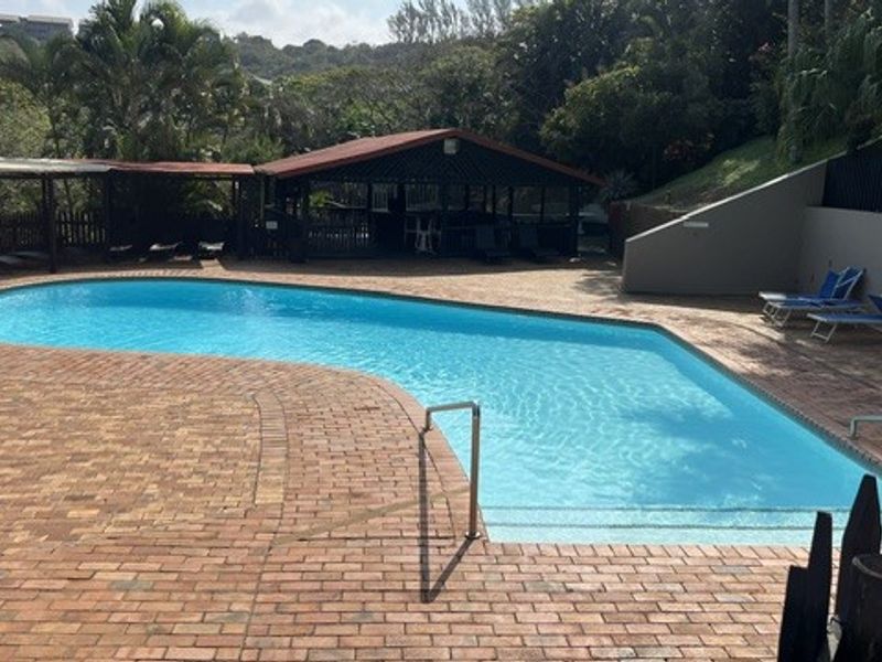A communal swimming pool area featuring brick paving, a covered gazebo, and a retaining wall, set within a landscaped outdoor environment.