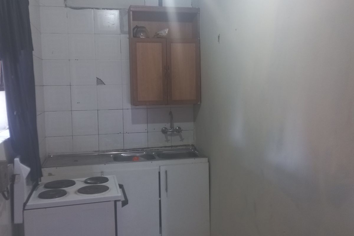 A compact kitchen area featuring a stainless steel sink, wall-mounted cabinetry, and a tiled splashback, currently showing signs of wear and dated finishes.