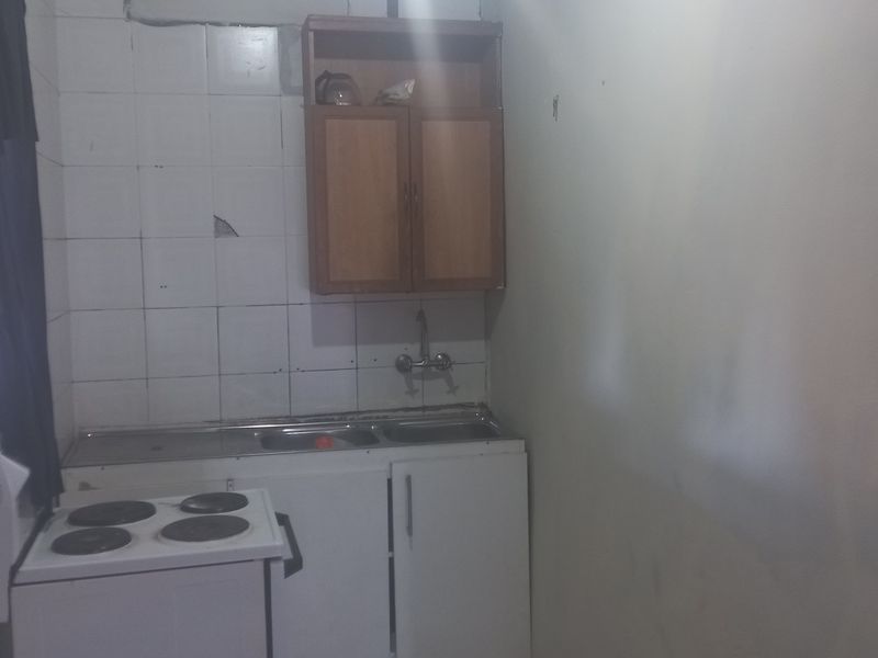 A compact kitchen area featuring a stainless steel sink, wall-mounted cabinetry, and a tiled splashback, currently showing signs of wear and dated finishes.