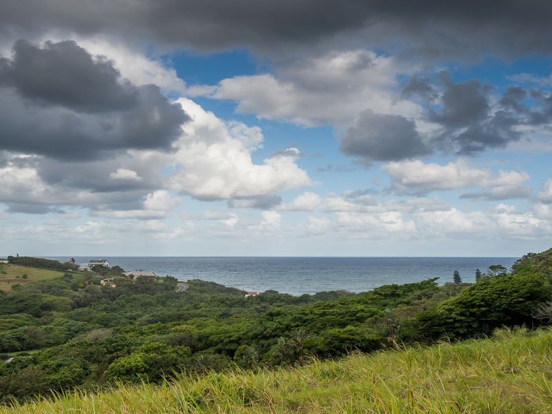 A scenic coastal landscape view featuring lush green vegetation, rolling hills, and an expansive ocean horizon under a cloudy sky.