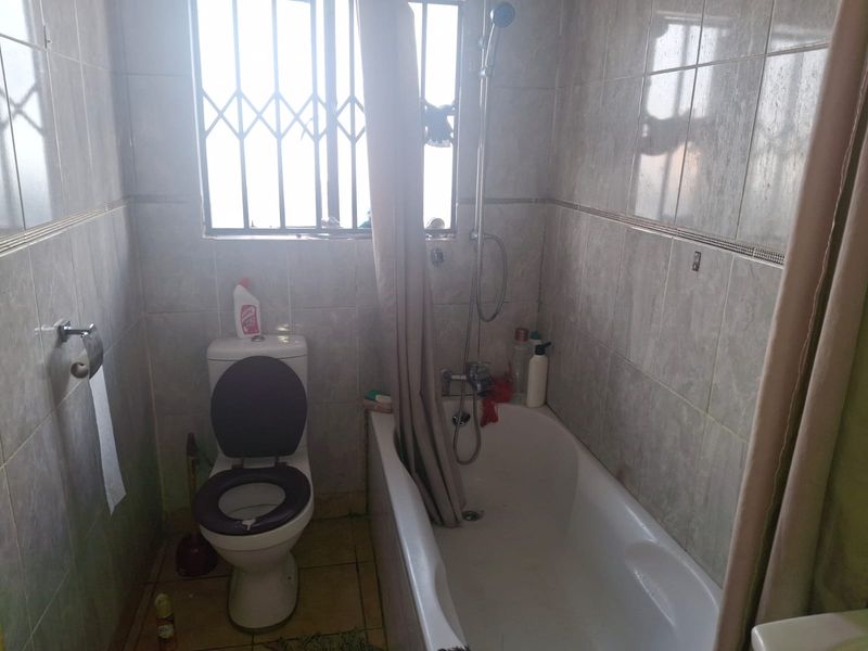 A compact bathroom featuring a bathtub with shower attachment, toilet, and full-height wall tiling with security bars on the window.
