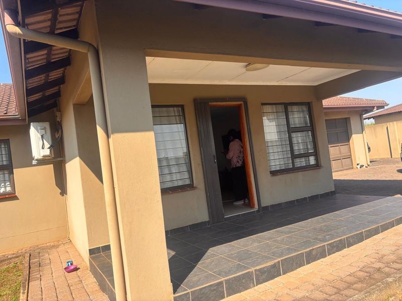 Front view of the townhouse featuring a covered tiled patio, paved driveway, and exterior wall-mounted air conditioning unit.