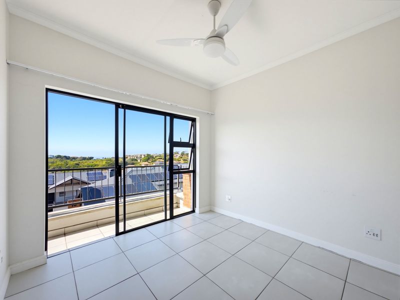 A bright, empty bedroom featuring tiled flooring, a ceiling fan, and large sliding glass doors opening onto a balcony with scenic views.