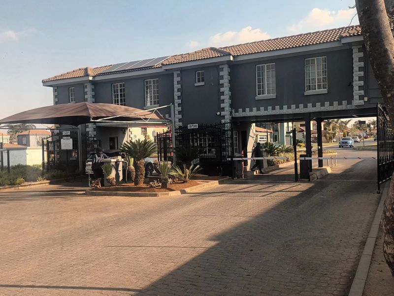 The entrance to the apartment complex features a secure gatehouse, boom gate access, and a paved driveway with solar panels installed on the roof.
