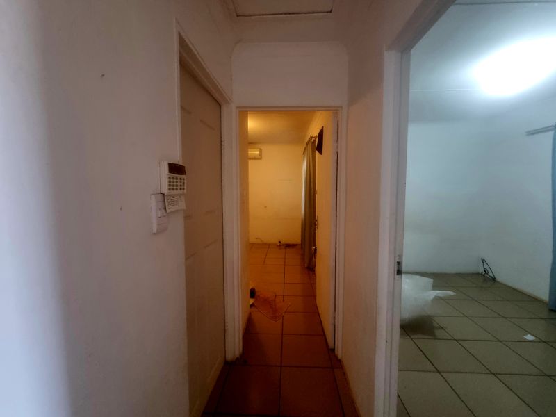 A narrow hallway with tiled flooring, connecting multiple rooms, featuring a wall-mounted security alarm keypad and basic lighting.