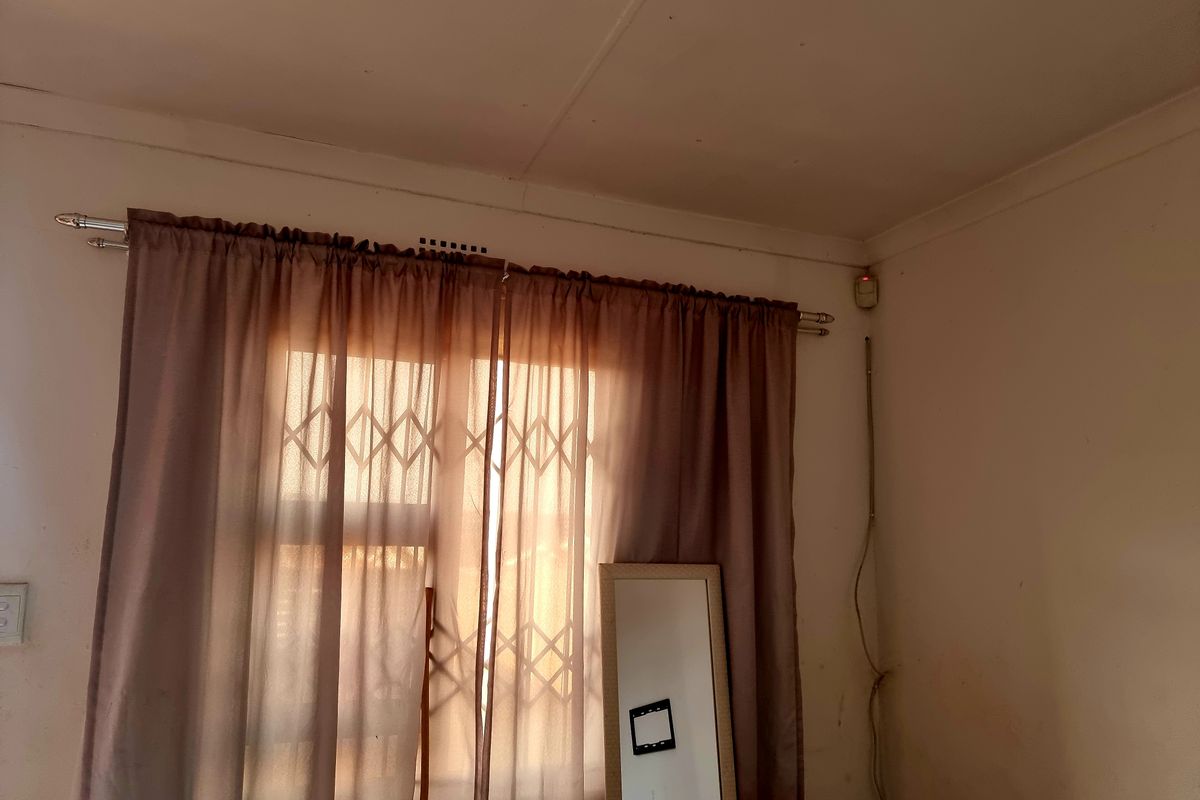 A bedroom interior featuring a window with a security gate, curtain rod, and wall-mounted alarm sensor.