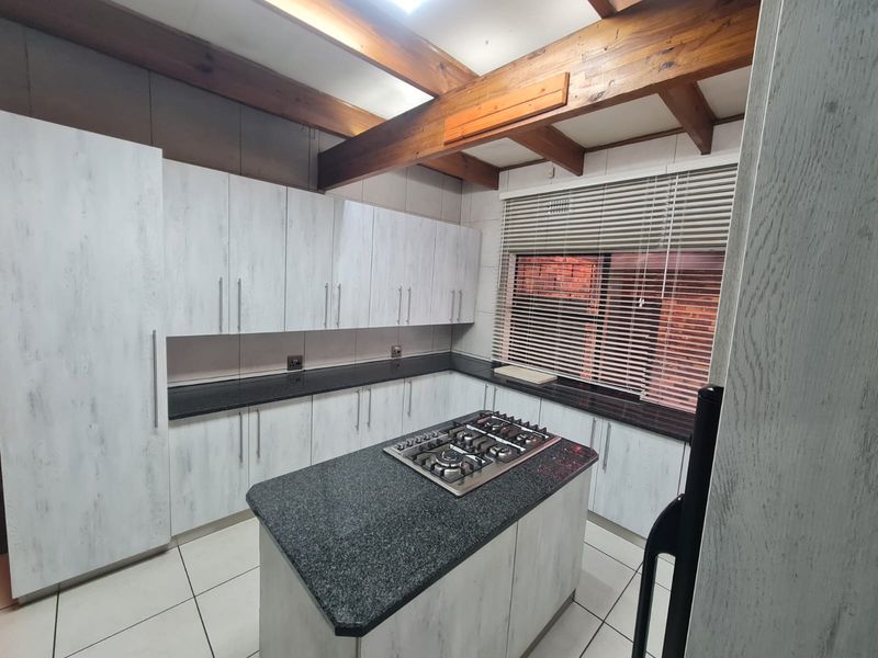 Modern kitchen featuring light wood-grain cabinetry, granite countertops, a central island with a gas hob, and exposed timber ceiling beams.