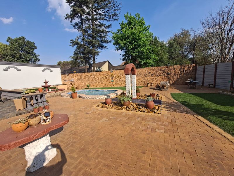A well-maintained outdoor entertainment area featuring a swimming pool, paved patio, decorative archway, and a combination of artificial lawn and brick boundary walls.
