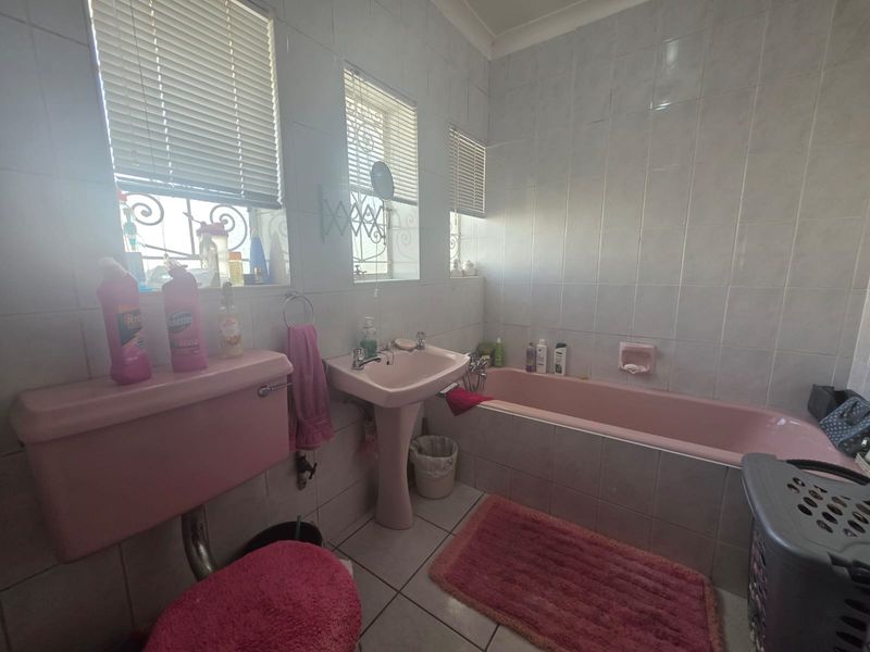 A dated bathroom featuring a pink pedestal basin, matching bathtub, and toilet cistern, with full-height wall tiling and security bars on the windows.