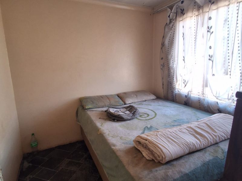 A compact bedroom featuring dark tiled flooring and a window with curtain rails, currently requiring ceiling repairs and general maintenance.