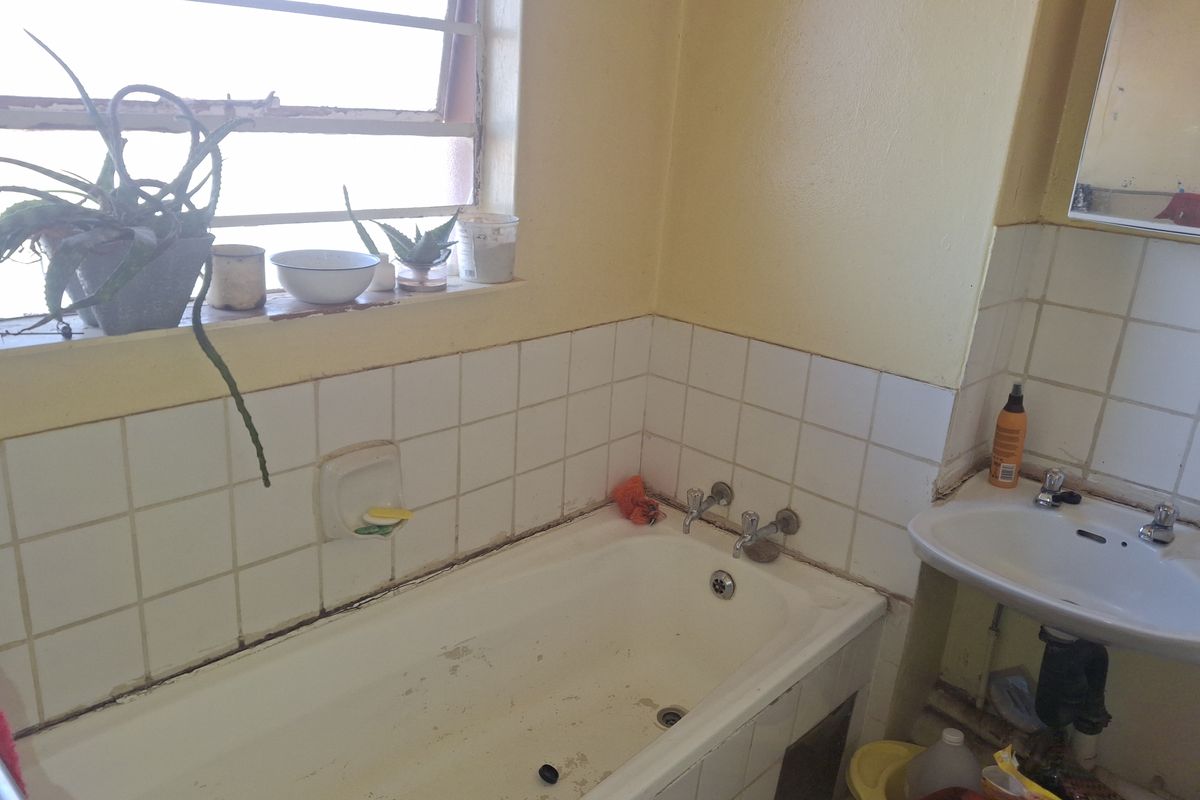 A dated bathroom featuring a built-in bathtub, wall-mounted basin, and louvered window, requiring renovation to update the finishes and address wear.