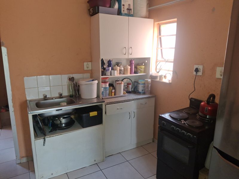 A compact kitchen featuring built-in cabinetry, a stainless steel sink, tiled splashback, and ceramic floor tiles, requiring cosmetic updates to improve its overall condition.