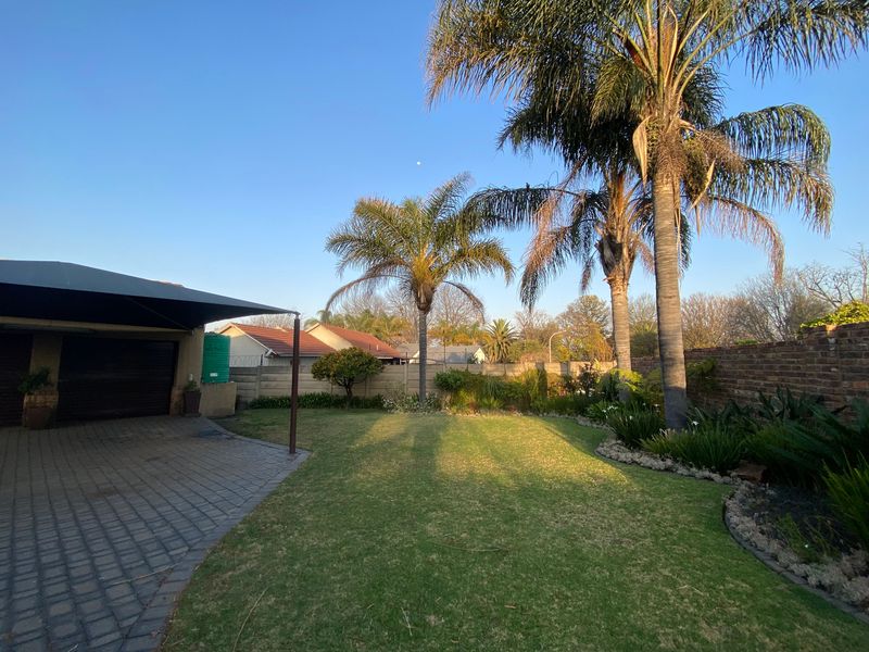 A well-maintained garden featuring a paved driveway, carport, established palm trees, and a secure brick boundary wall.