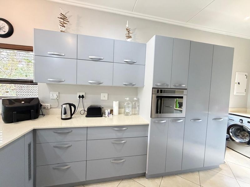A modern kitchen featuring light grey cabinetry, stone countertops, a built-in oven, and an integrated laundry space with tiled flooring.