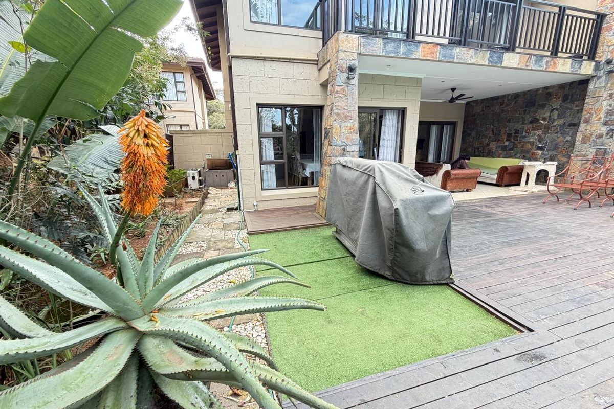 Rear exterior view of a townhouse featuring a covered patio, timber decking, artificial lawn, and stone-clad facade with an upper-level balcony.