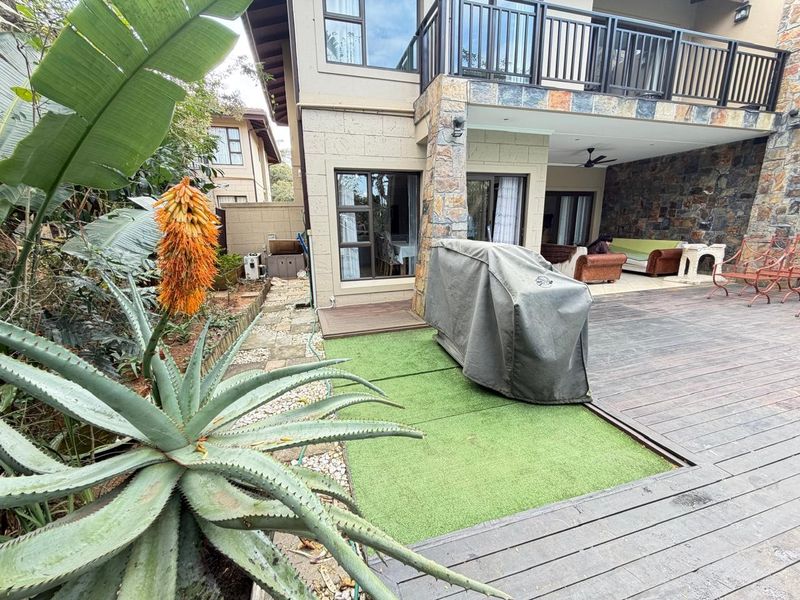 Rear exterior view of a townhouse featuring a covered patio, timber decking, artificial lawn, and stone-clad facade with an upper-level balcony.