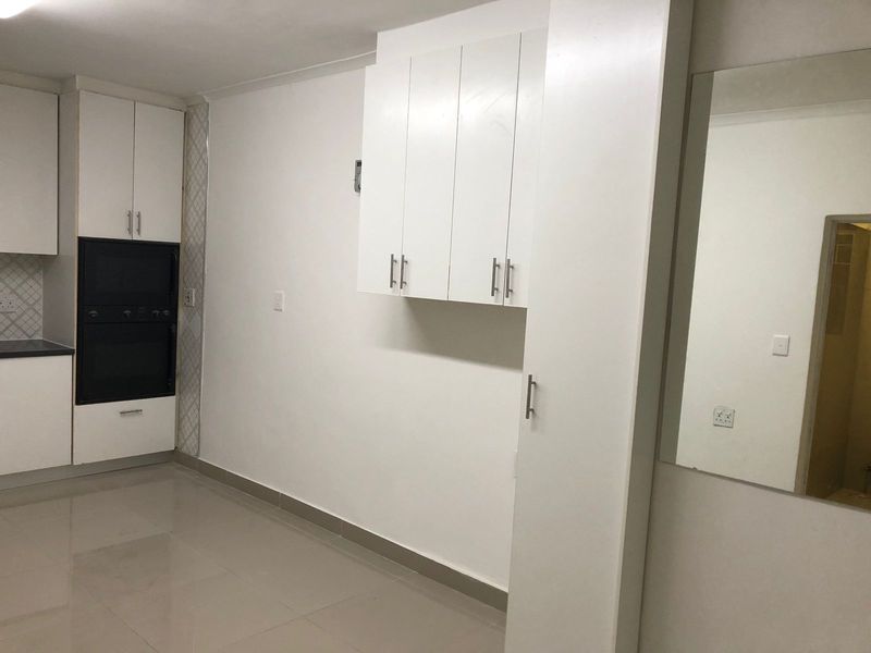 A clean, modern kitchen area featuring white built-in cabinetry, integrated oven and microwave, and neutral tiled flooring.