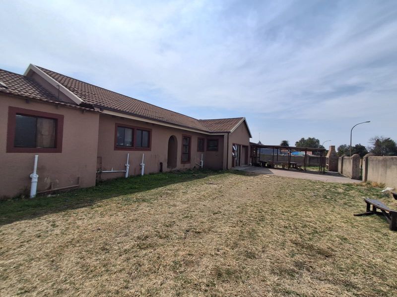 Rear view of the property featuring a tiled roof, a covered patio area with a built-in braai, and a large backyard with boundary walls.