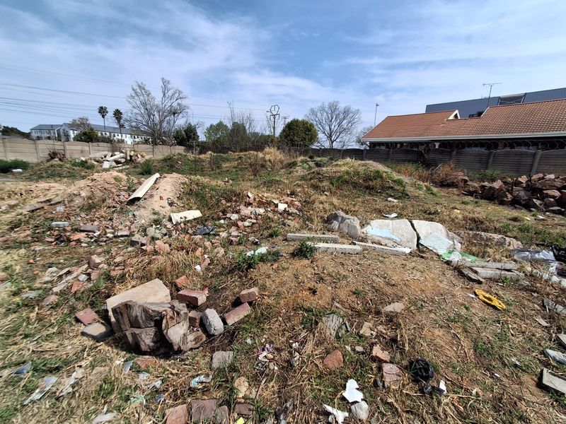 An undeveloped plot of land featuring uneven terrain, scattered rubble, and debris, enclosed by a perimeter wall with security fencing.