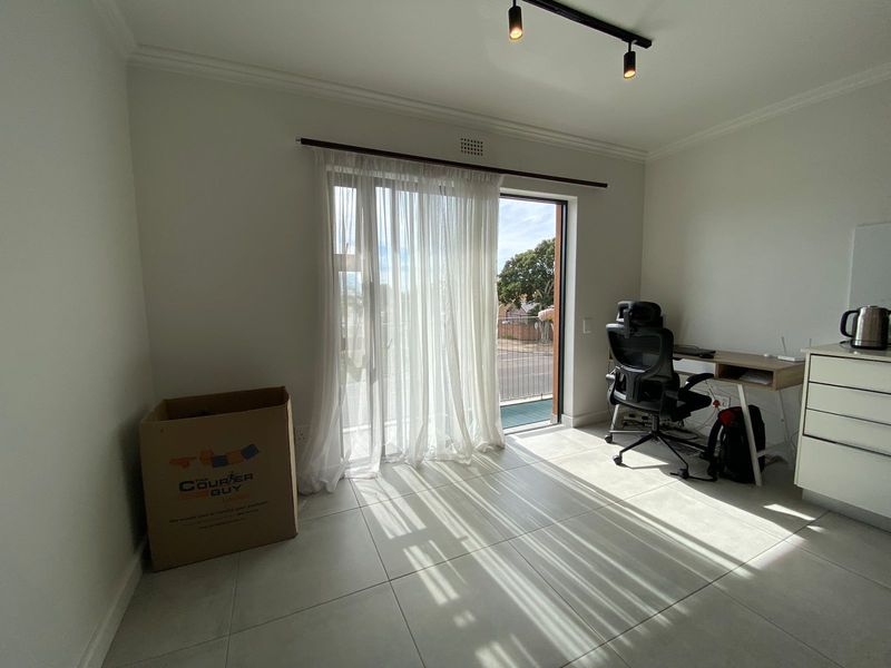 A bright bedroom featuring large sliding glass doors that open to a balcony, neutral tiled flooring, and modern track lighting.