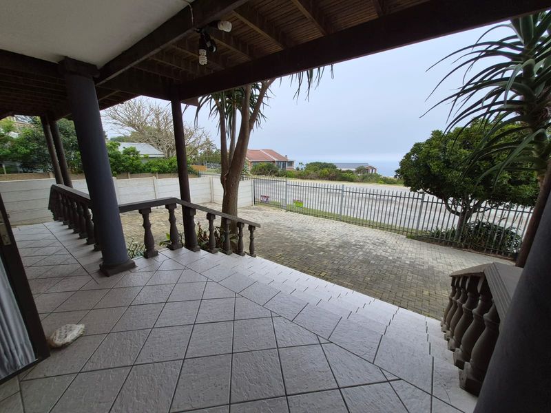 A covered patio area featuring tiled flooring and a timber ceiling, overlooking a paved driveway and perimeter fencing with a distant sea view.