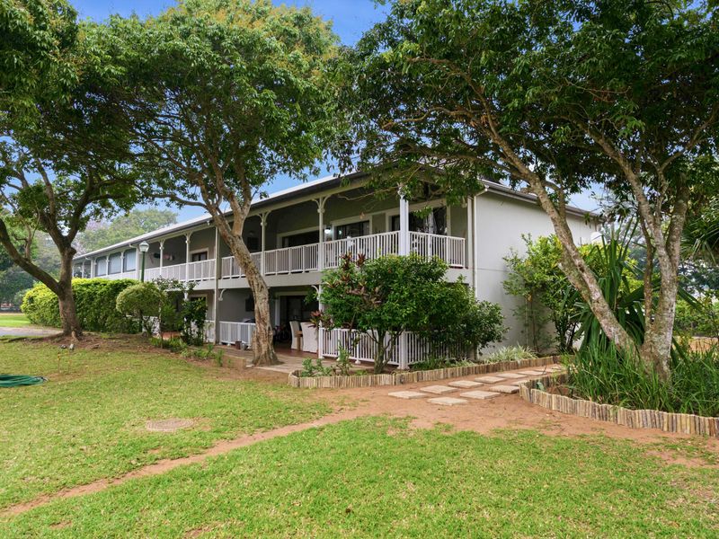 Rear view of the apartment building featuring a multi-level balcony with white railings, surrounded by mature trees and a landscaped garden area.