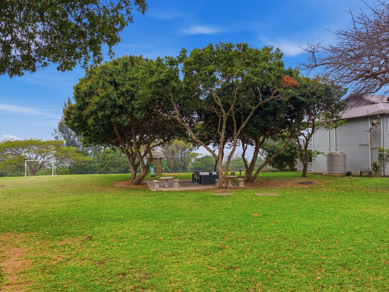A spacious communal garden area featuring mature trees, a paved seating space, and a water tank, adjacent to the apartment building.