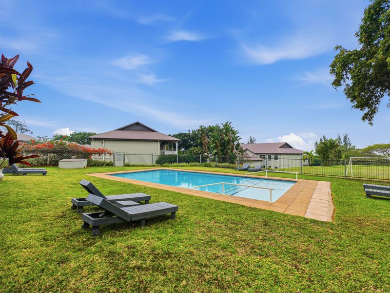 Outdoor communal pool area featuring a rectangular swimming pool with safety railing, paved surround, and surrounding lawn within a fenced complex.