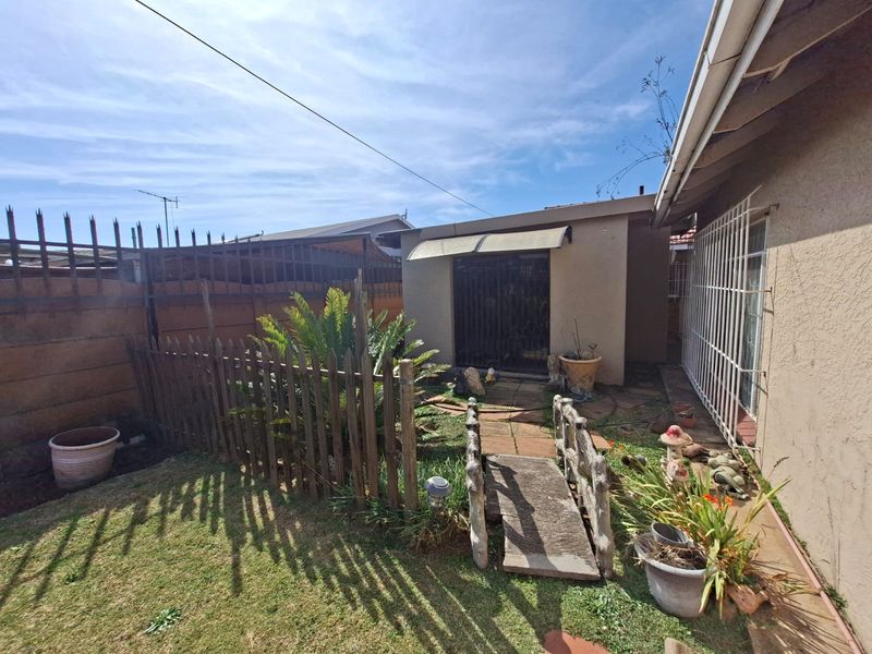 Rear exterior area featuring a small garden, paved walkway with a decorative bridge, security fencing, and a covered entrance to the main dwelling.