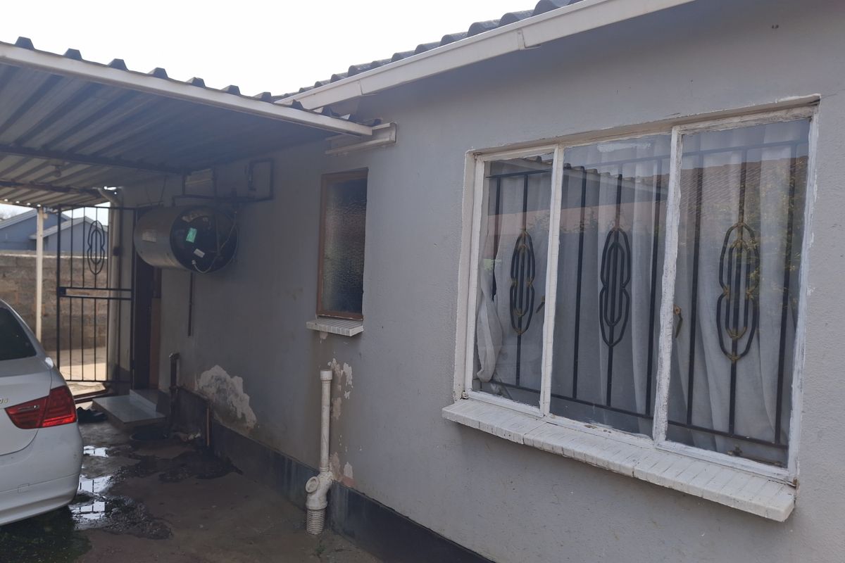 Rear exterior view of the house featuring a carport, external geyser, and windows with security bars, showing signs of exterior wall maintenance required.