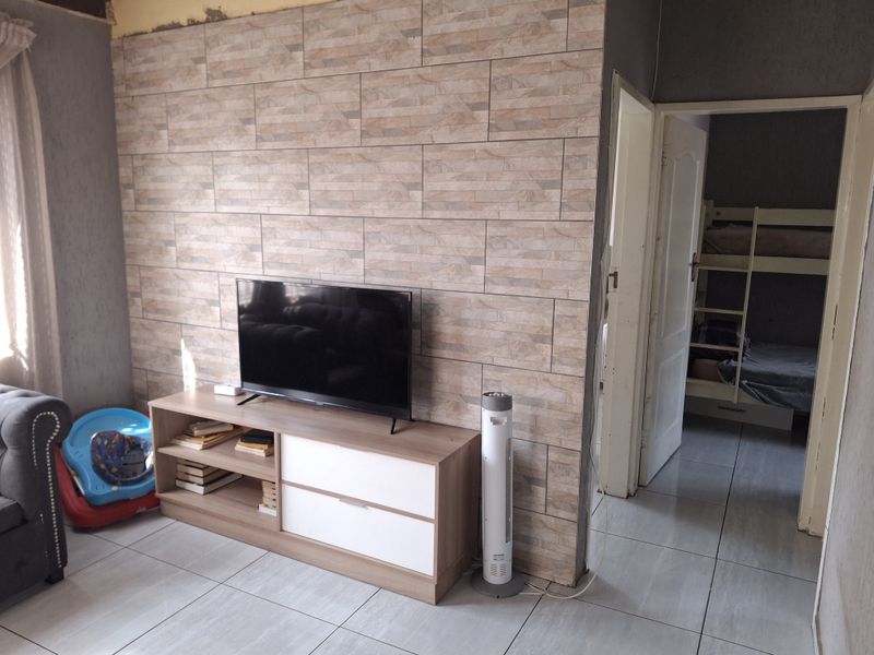 Living room area featuring a tiled accent wall and ceramic floor tiles, with an adjacent bedroom visible through the doorway.