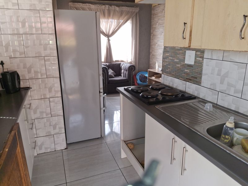 A compact kitchen featuring tiled splashbacks, a built-in electric hob, and stainless steel sink, with some cabinetry requiring maintenance or replacement.