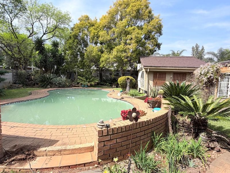 Outdoor pool area featuring a curved brick retaining wall, paved surround, and an adjacent outbuilding set within a mature garden.