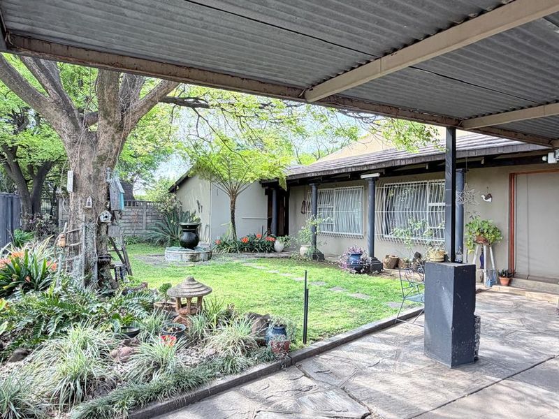 A covered patio area overlooking a landscaped garden with mature trees and paved pathways, featuring a corrugated metal roof structure.